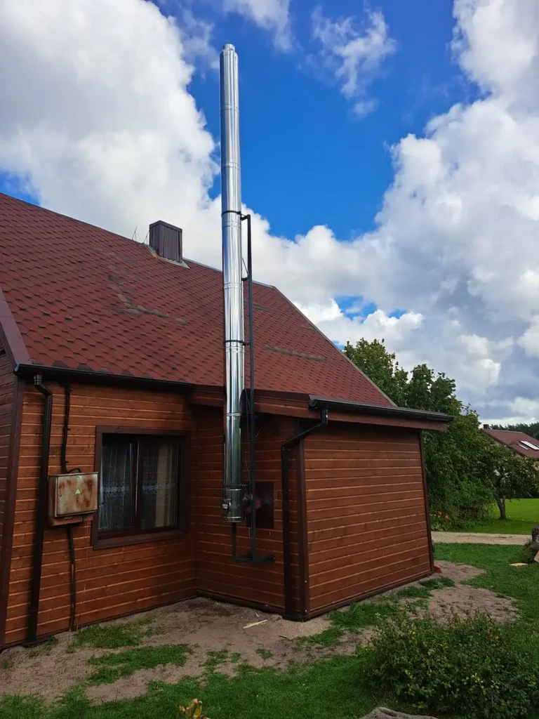 Raudonas plytų pastatas su rudu stogu ir aukštu, sidabriniu kaminu. Dangus yra debesuotas pilkas.