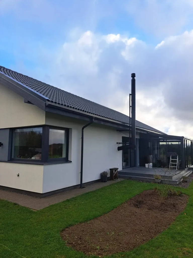 Baltas vieno aukšto namas su pilku stogu ir aukštu, sidabriniu kaminu. Namas turi langą kairėje pusėje ir mažą krūmą priekyje. Dangus yra debesuotas mėlynas.