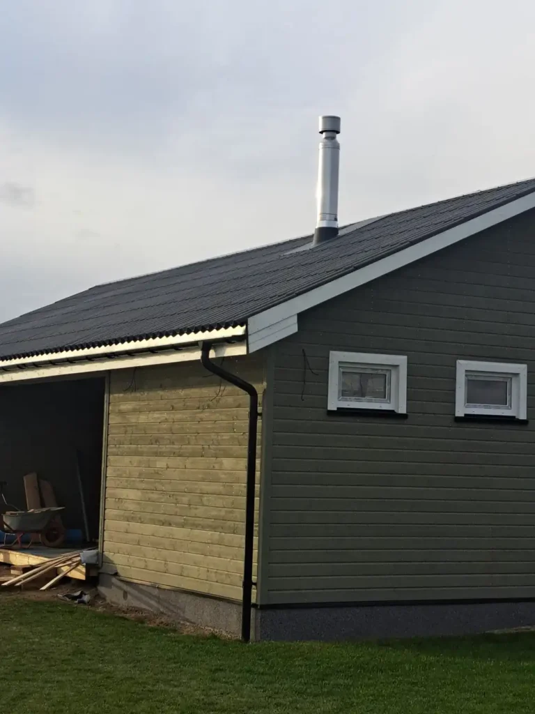 Tamsiai žalias namas su pilku stogu ir aukštu, sidabriniu kaminu. Namas turi medines duris ir langą šone. Fone matosi medžiai ir krūmai.